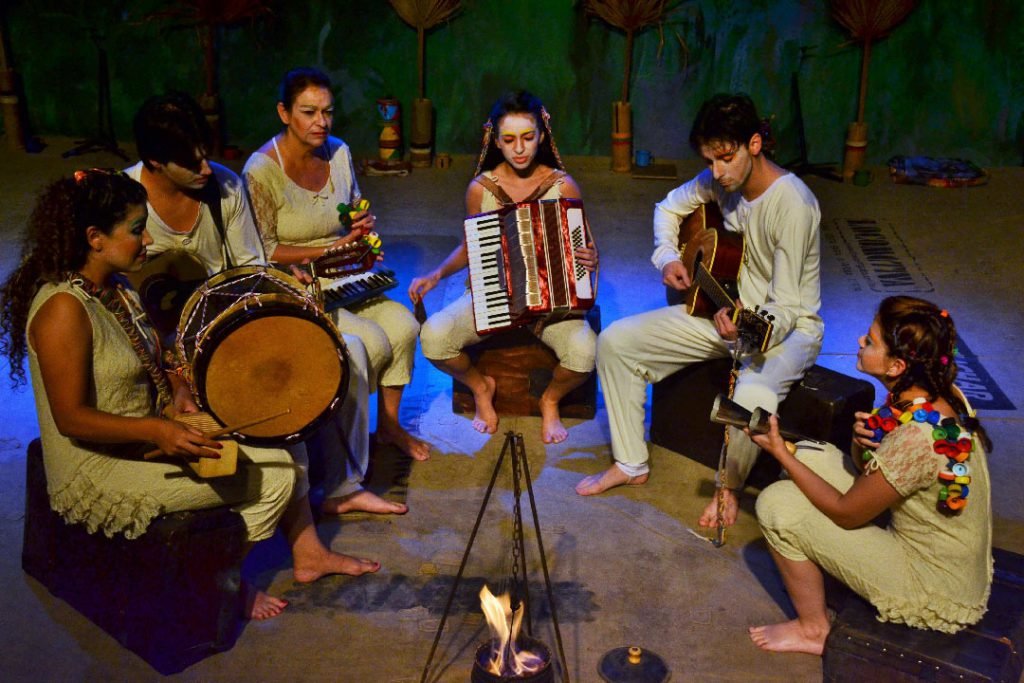 Seis atores descalços, de roupas cor de creme, sentados em roda ao redor de uma pequena chama, tocando bombo, sanfona, violão, teclado e reco-reco. Ao fundo, um cenário pintado de vegetação seca.