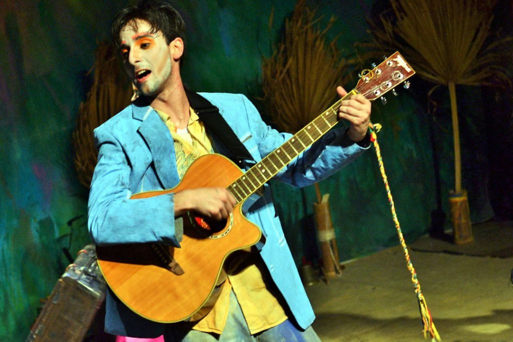 Um ator de rosto pintado, paletó azul-claro sobre camisa amarela, canta tocando violão diante do cenário de vegetação seca.