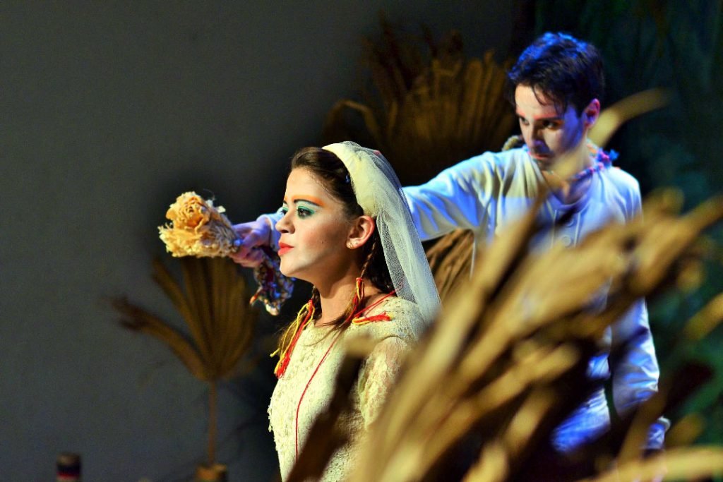 Uma atriz vestida de noiva, com véu curto e buquê de flores secas, olha para longe enquanto um ator de roupa branca ajeita o véu atrás dela.