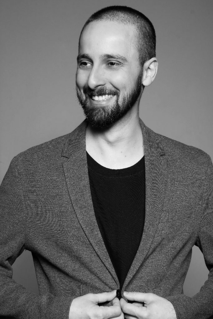 Retrato em preto e branco de Rafael Érnica, de barba e cabelo curto, sorrindo enquanto abotoa o paletó sobre uma camiseta preta.