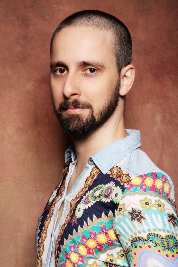 Retrato de Rafael Érnica de perfil parcial, sério, com camisa estampada de flores e gola jeans, diante de um fundo terroso.
