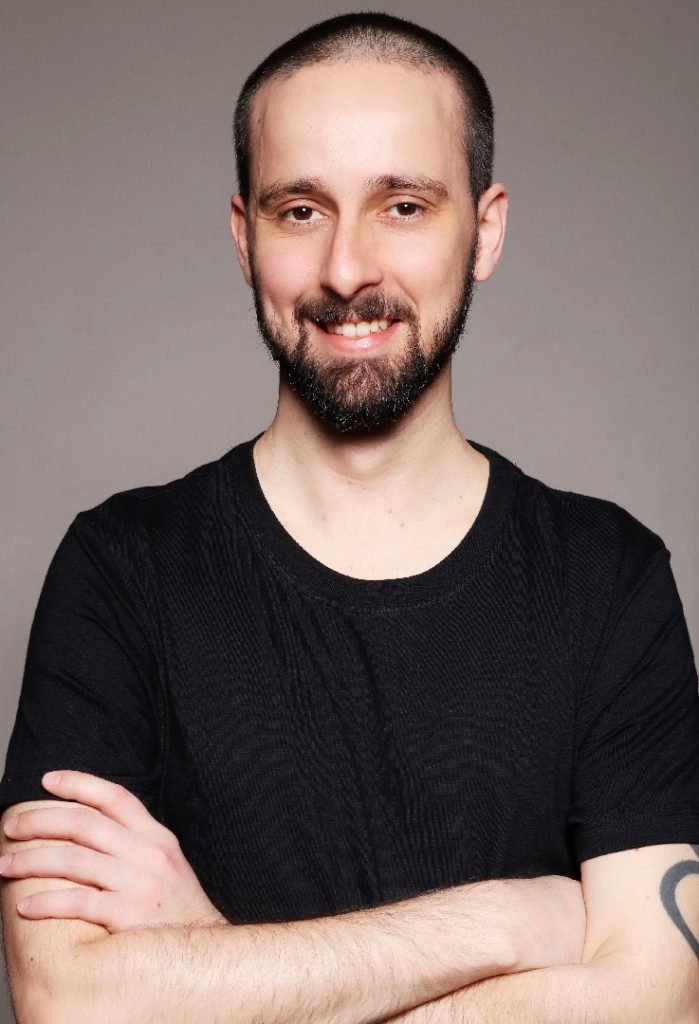 Retrato de Rafael Érnica de camiseta preta e braços cruzados, sorrindo para a câmera diante de um fundo cinza.