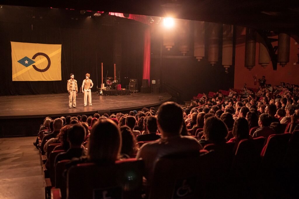 Vista do fundo de uma plateia lotada: dois atores em cena no palco, e à esquerda a bandeira amarela com losango azul e um círculo escuro. Ao fundo do palco, os instrumentos da banda.
