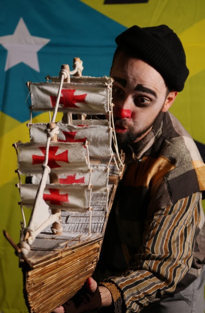 Um ator de nariz vermelho, gorro preto e jaqueta de retalhos listrados segura uma caravela de madeira cujas velas trazem cruzes vermelhas de cruzada; ao fundo, a bandeira amarela e azul com a estrela branca.