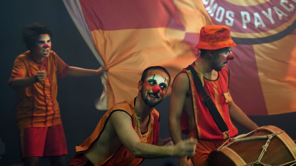 Três atores de nariz vermelho e roupa laranja: um toca um bombo, outro puxa um pano listrado de laranja e creme; ao fundo, uma bandeira com a palavra “payaços”.