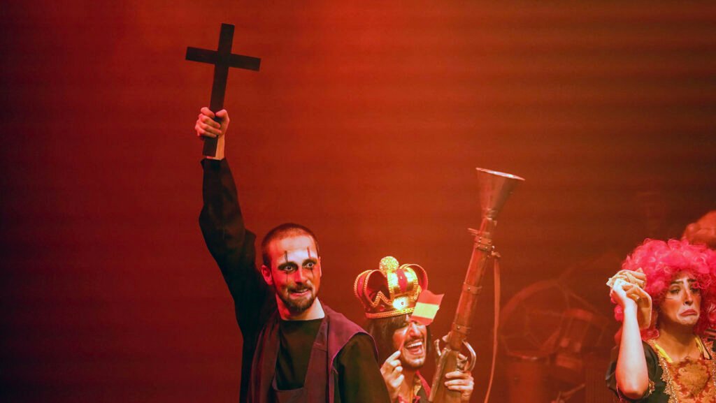Sob luz vermelha, um ator de manto escuro e rosto pintado ergue uma cruz de madeira; ao lado, uma figura de coroa dourada ri segurando uma arma em forma de corneta, e à direita outra, de peruca rosa, faz careta de choro.