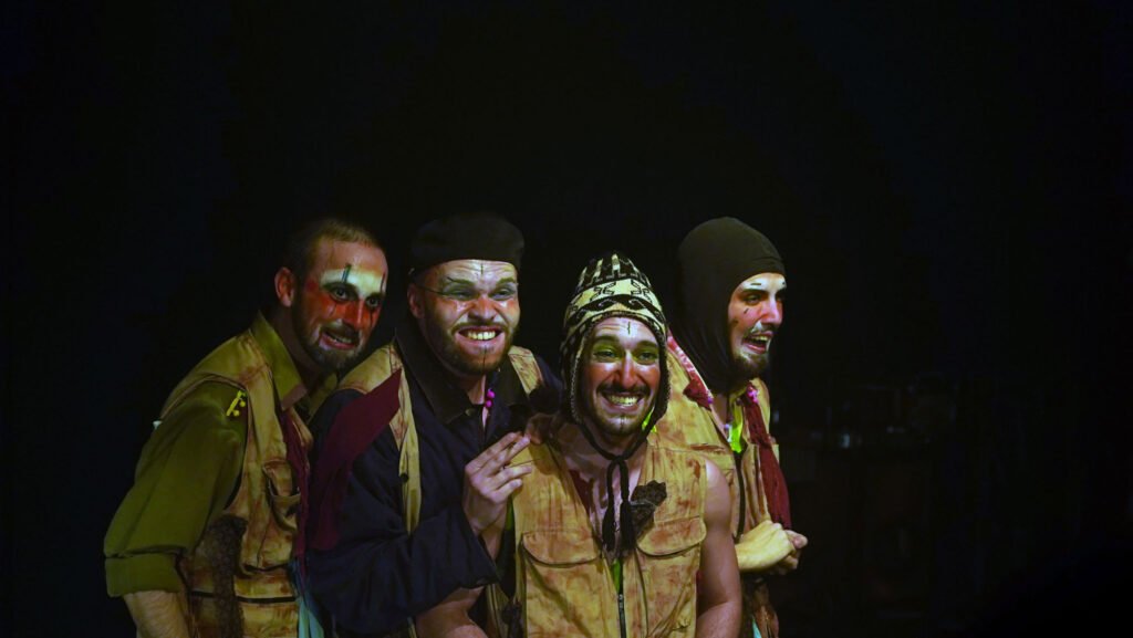 Quatro atores de ¡CERRADO! lado a lado, rostos pintados de branco e laranja, em coletes e jaquetas de tom terroso; um usa gorro andino, outro uma balaclava preta. Sorriem para a plateia no escuro.