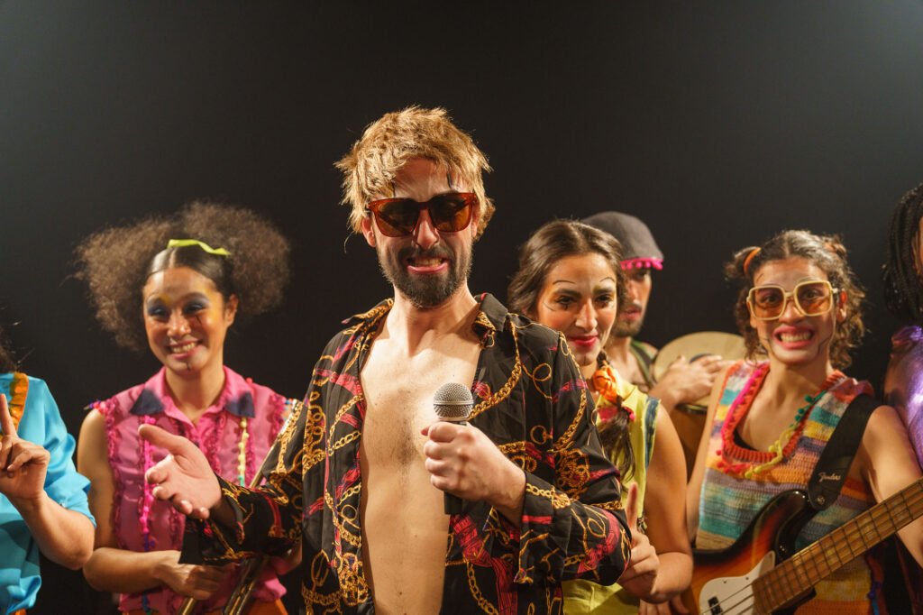 Um ator de peruca loira, óculos escuros e camisa aberta estampada de correntes segura o microfone, cercado pelo elenco em roupas coloridas, com baixo e pandeiro nas mãos.