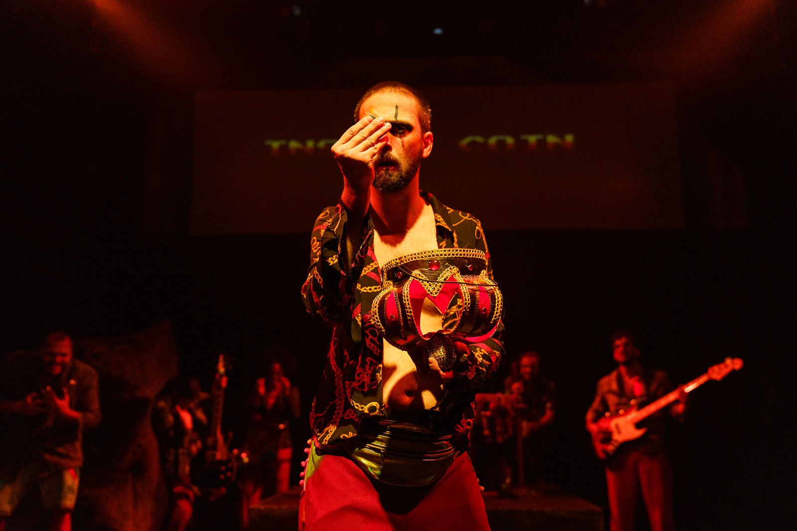 Cena de ¡CERRADO!: sob luz vermelha, um ator de camisa estampada aberta segura diante do peito um adereço dourado em forma de coroa e cobre um dos olhos com a mão; atrás dele, músicos com guitarra e baixo.