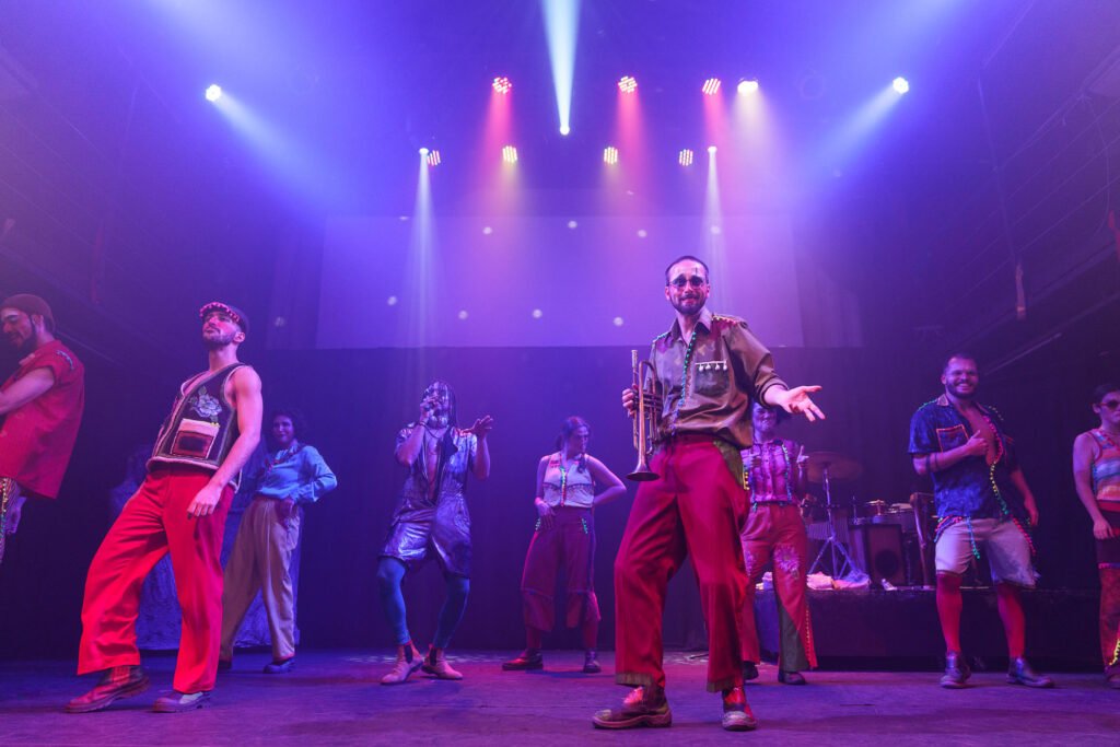O elenco inteiro em cena sob luz violeta: ao centro, um ator de camisa verde-oliva e calça vermelha estende o braço segurando um trompete; os demais dançam, um canta ao microfone, e há bateria à direita.