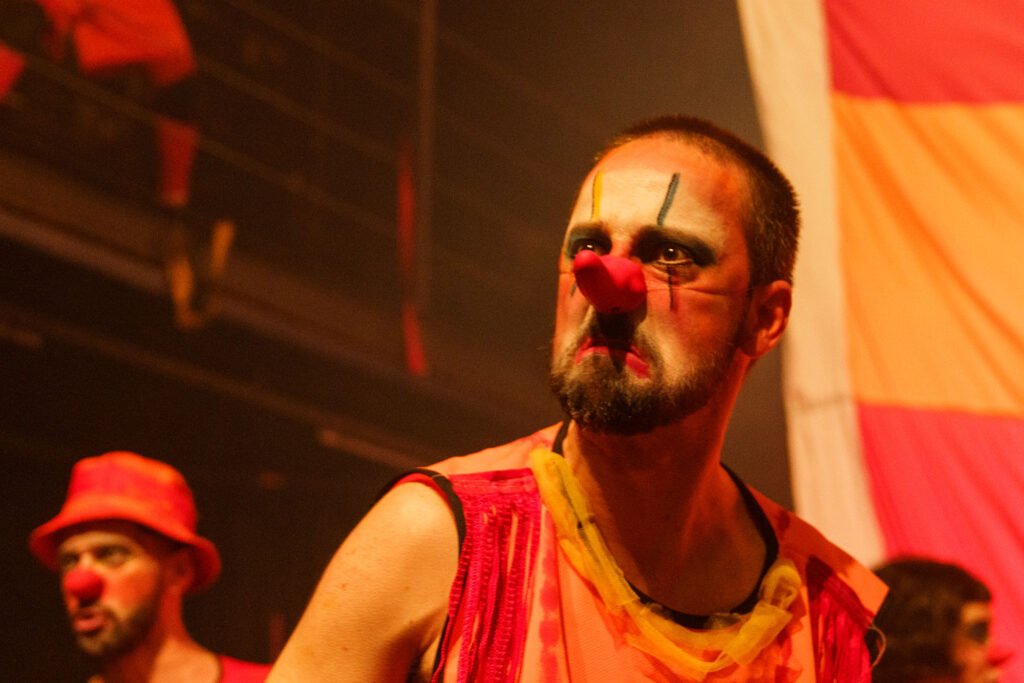 Retrato de um palhaço de ¡CERRADO!: rosto branco, nariz vermelho, sobrancelhas pintadas e barba, em colete rosa e amarelo, com expressão séria. Atrás, outro palhaço de chapéu vermelho.