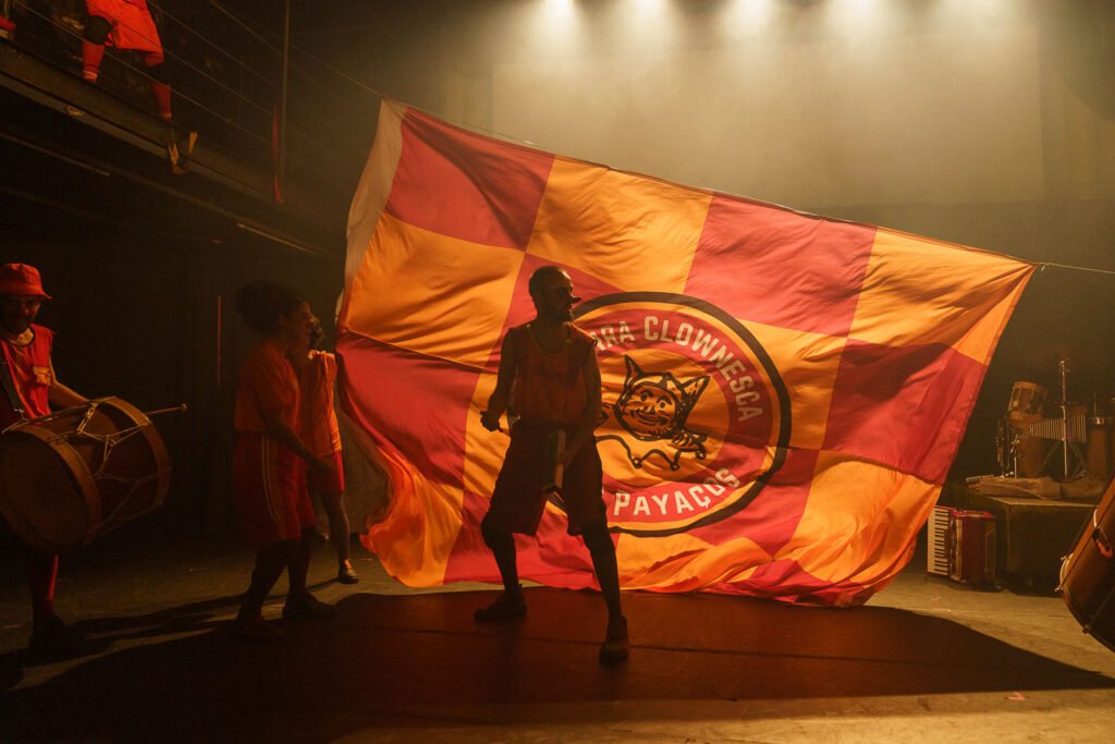 Uma bandeira enorme de quadrados vermelhos e laranja atravessa o palco, com o emblema de um bufão e os dizeres “clownesca” e “payaços”. Atores de vermelho a seguram; à esquerda, um bombo.